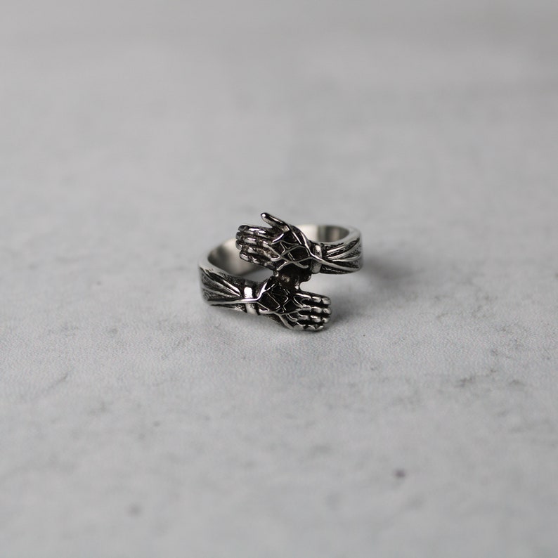 May include: A silver ring with two hands clasped together, forming a circle. The ring is made of metal and has a detailed design.