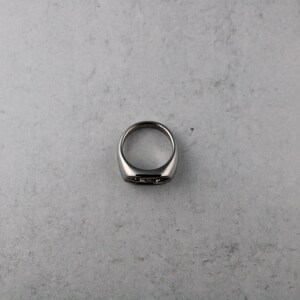 May include: A silver-colored ring with a flat, rectangular face. The ring has a smooth, polished finish and a thick band. The face of the ring has a subtle, dark design. The ring is shown on a light gray surface.