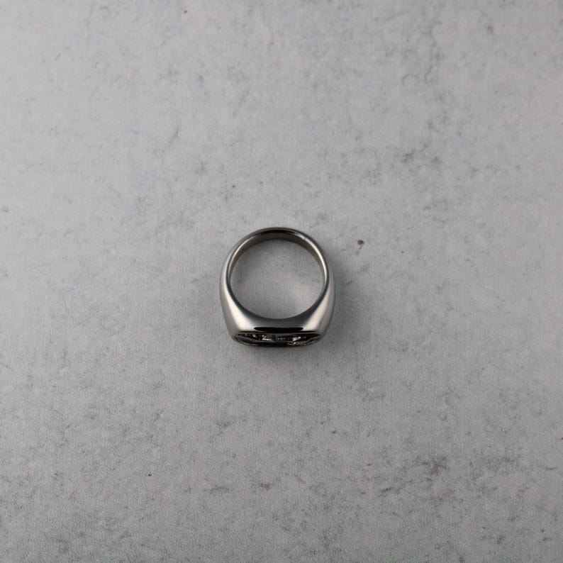 May include: A silver-colored ring with a flat, rectangular face. The ring has a smooth, polished finish and a thick band. The face of the ring has a subtle, dark design. The ring is shown on a light gray surface.