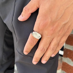 May include: A silver ring with a rectangular face featuring a detailed design. The ring is worn on a finger, with the hand and part of a dark-colored outfit visible in the image. The ring's design appears to be a landscape or abstract pattern.