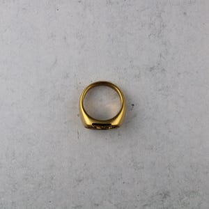 May include: A gold-colored signet ring with a smooth, polished surface. The ring has a rounded shape and a flat top with a subtle engraved design. The ring is set against a neutral background, highlighting its metallic sheen.