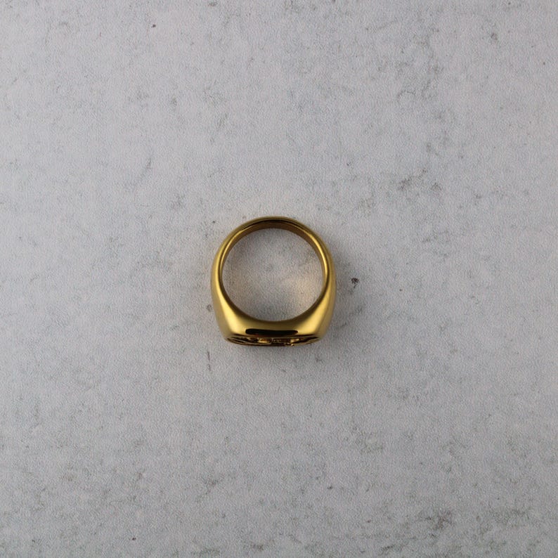 May include: A gold-colored signet ring with a smooth, polished surface. The ring has a rounded shape and a flat top with a subtle engraved design. The ring is set against a neutral background, highlighting its metallic sheen.