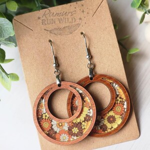 May include: Wooden hoop earrings with a retro floral pattern in shades of orange, yellow, and white. The earrings are on a brown card with the text "Rumurs Run Wild". The earrings have silver-colored hooks.