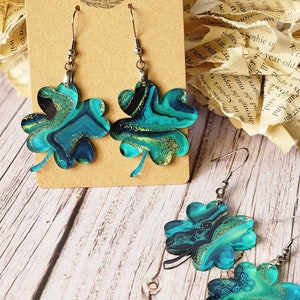 May include: A pair of earrings featuring four-leaf clover charms with a blue, green, and gold marbled design. The earrings are on a brown card with the text "Rumurs RUN WILD".