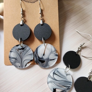 May include: A pair of dangle earrings with black and white marbled round resin pieces. The earrings have silver hooks and a black round resin piece above the marbled piece. The earrings are displayed on a brown card with the text "Rumors Run Wild" printed on it.