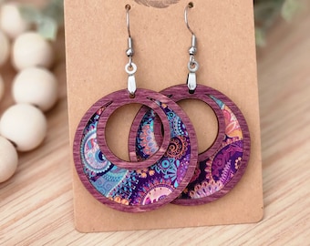 May include: A pair of hoop earrings with a purple wooden frame and a colorful paisley design. The earrings are hanging on a brown card with the text "Rumurs Run Wild". The earrings have silver-colored hooks.