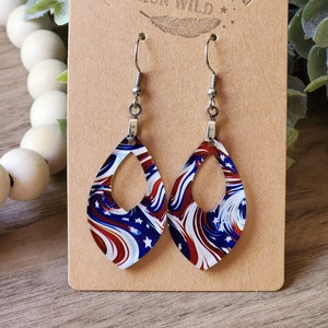 May include: A pair of dangle earrings with a red, white, and blue swirl pattern. The earrings are shaped like a teardrop with a cutout in the center. The earrings are hanging on a brown card with the text "Rumors Run Wild" printed on it.