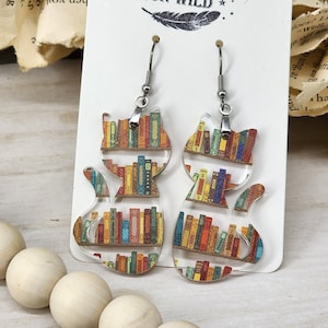 May include: A pair of cat-shaped earrings with a bookshelf design. The earrings are made of clear acrylic and feature a colorful pattern of books on shelves. The earrings are hanging from silver hooks.