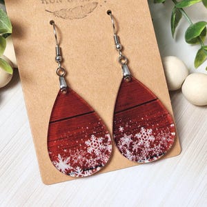 May include: A pair of teardrop-shaped earrings with a festive winter design. The earrings feature a red wood-grain background with white snowflakes. The earrings are displayed on a brown card with the text "Rumurs Run Wild".