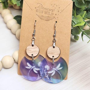 May include: A pair of dangle earrings with a nature theme. Each earring features a wooden circle, a silver-tone hook, and a translucent, half-circle pendant with a dragonfly and floral design. The pendant has purple, blue, and green hues. The earrings are displayed on a brown card.