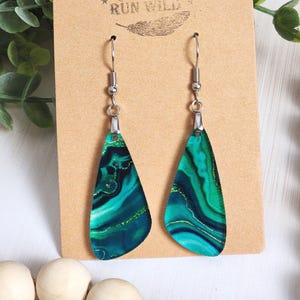 May include: A pair of dangle earrings with a malachite design. The earrings have a teardrop shape with a green and blue marbled pattern and gold accents. The earrings hang from silver-colored hooks and are displayed on a brown card with the text "Rumurs RUN WILD".