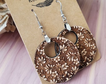 May include: A pair of wooden earrings with a floral pattern. The earrings are round and have a hole in the center. The earrings are hanging from silver hooks. The earrings are on a brown card with the text "Rumors Run Wild" printed on it.