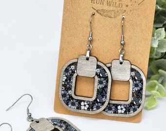 May include: A pair of silver earrings with a floral design. The earrings are made of wood and have a square shape with a cutout in the center. The floral design is in shades of blue and white.