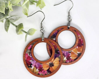 May include: A pair of wooden earrings with a floral pattern. The earrings are brown with a pink and yellow floral design. The earrings are in the shape of a circle with a smaller circle cut out of the center.