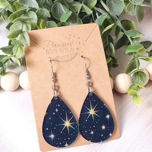 May include: Teardrop-shaped earrings with a dark blue background and a celestial star pattern. The earrings are suspended from silver-colored hooks and are displayed on a brown card with the text "Rumurs Run Wild".