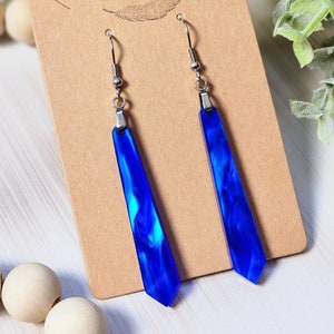 Peut inclure: Une paire de boucles d'oreilles pendantes bleu et argent avec un effet marbré. Les boucles d'oreilles sont sur une carte marron avec le texte "Rumors Run Wild" et un motif de plume.