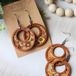 May include: Wooden hoop earrings with a retro floral design in shades of orange, yellow, and brown. The earrings have a double-hoop design with a floral pattern inside the hoops. The earrings are displayed on a brown card.