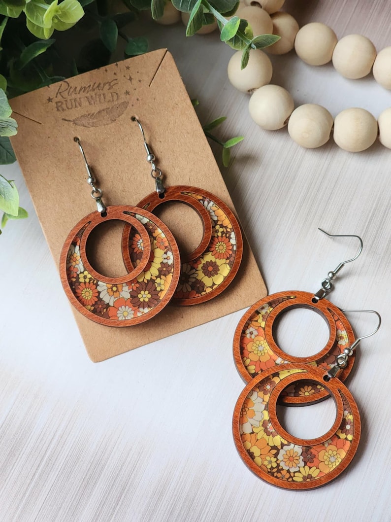 May include: Wooden hoop earrings with a retro floral design in shades of orange, yellow, and brown. The earrings have a double-hoop design with a floral pattern inside the hoops. The earrings are displayed on a brown card.