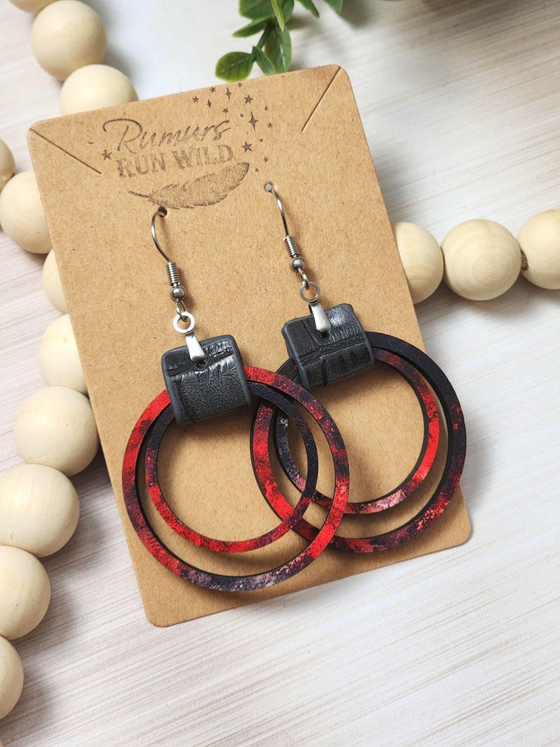 Puede incluir: Un par de pendientes colgantes con la parte superior negra similar al cuero y aros estampados en rojo y negro. Los pendientes cuelgan de ganchos plateados. Los pendientes se exhiben en una tarjeta marr&oacute;n con el texto "Rumurs Run Wild".