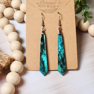 May include: Teal and black resin earrings with a marbled effect. The earrings are on a brown card with the text "Rumors Run Wild" printed on it.
