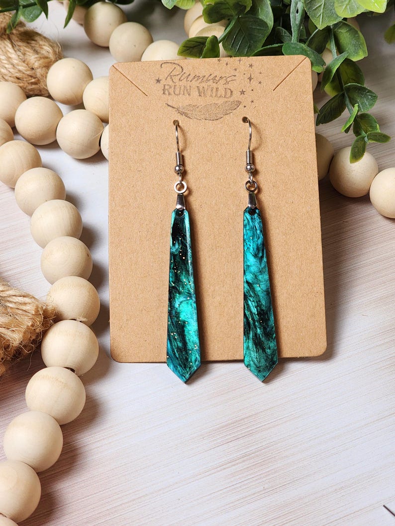 May include: Teal and black resin earrings with a marbled effect. The earrings are on a brown card with the text "Rumors Run Wild" printed on it.