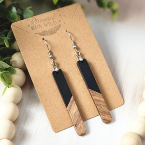 May include: A pair of rectangular earrings with a two-tone design. The top portion is black, and the bottom is a light brown wood grain pattern. The earrings hang from silver-colored hooks and are displayed on a brown card with the text "Rumors Run Wild".