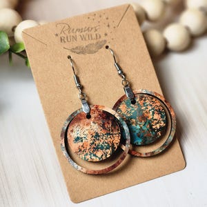 May include: A pair of circular earrings with a layered design. The earrings have a rusty orange and teal colour scheme with a circular outer frame. They are displayed on a brown card with the text "Rumurs Run Wild".