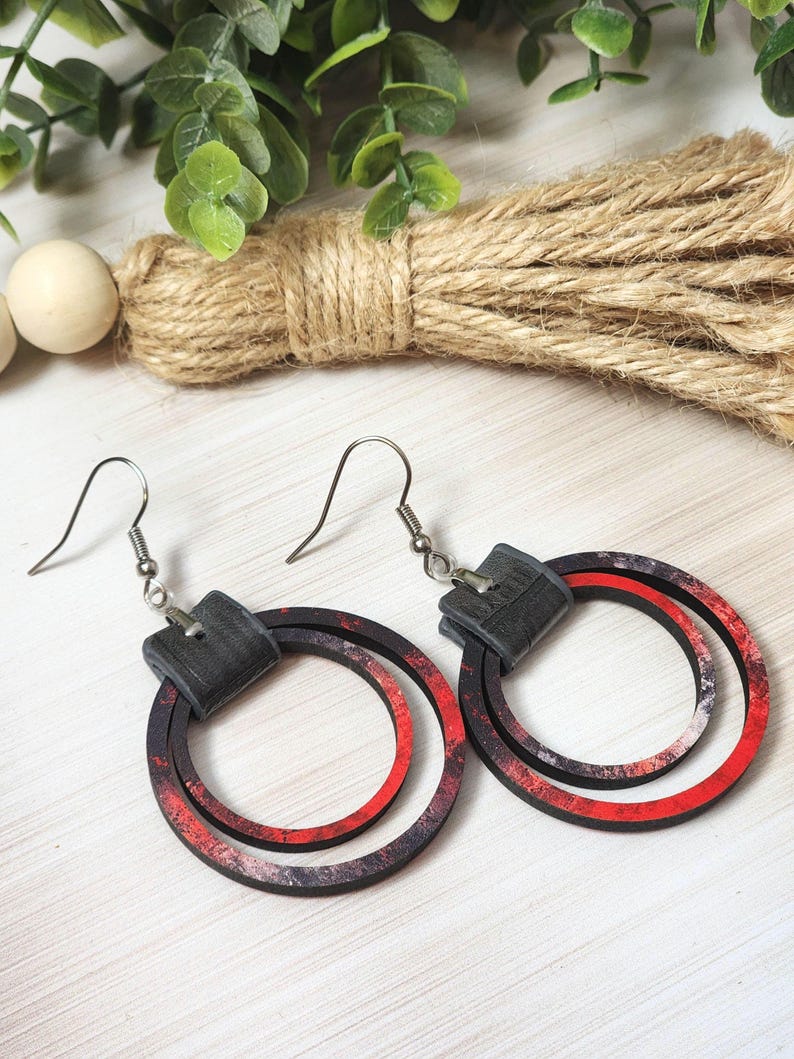 Puede incluir: Un par de pendientes de aro con un dise&ntilde;o en capas. Los pendientes presentan una parte superior cuadrada negra y dos c&iacute;rculos conc&eacute;ntricos rojos y negros. Los pendientes son de madera y tienen ganchos plateados.