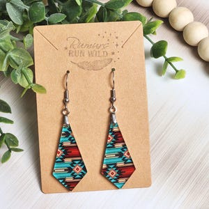 May include: A pair of diamond-shaped earrings with a Southwestern pattern in turquoise, red, and cream colors. The earrings hang from silver-colored hooks and are displayed on a brown card with the text "Rumurs Run Wild".