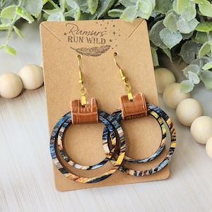 May include: A pair of wooden hoop earrings with a brown leather accent. The hoops are layered and have a blue, yellow, and brown pattern. The earrings are on a brown card with the text "Rumors Run Wild" printed on it.