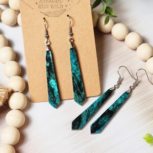 May include: A pair of long, teal and black dangle earrings with a silver hook. The earrings are made of resin and have a marbled effect. The earrings are on a brown card with the text "Rumours Run Wild" printed on it.
