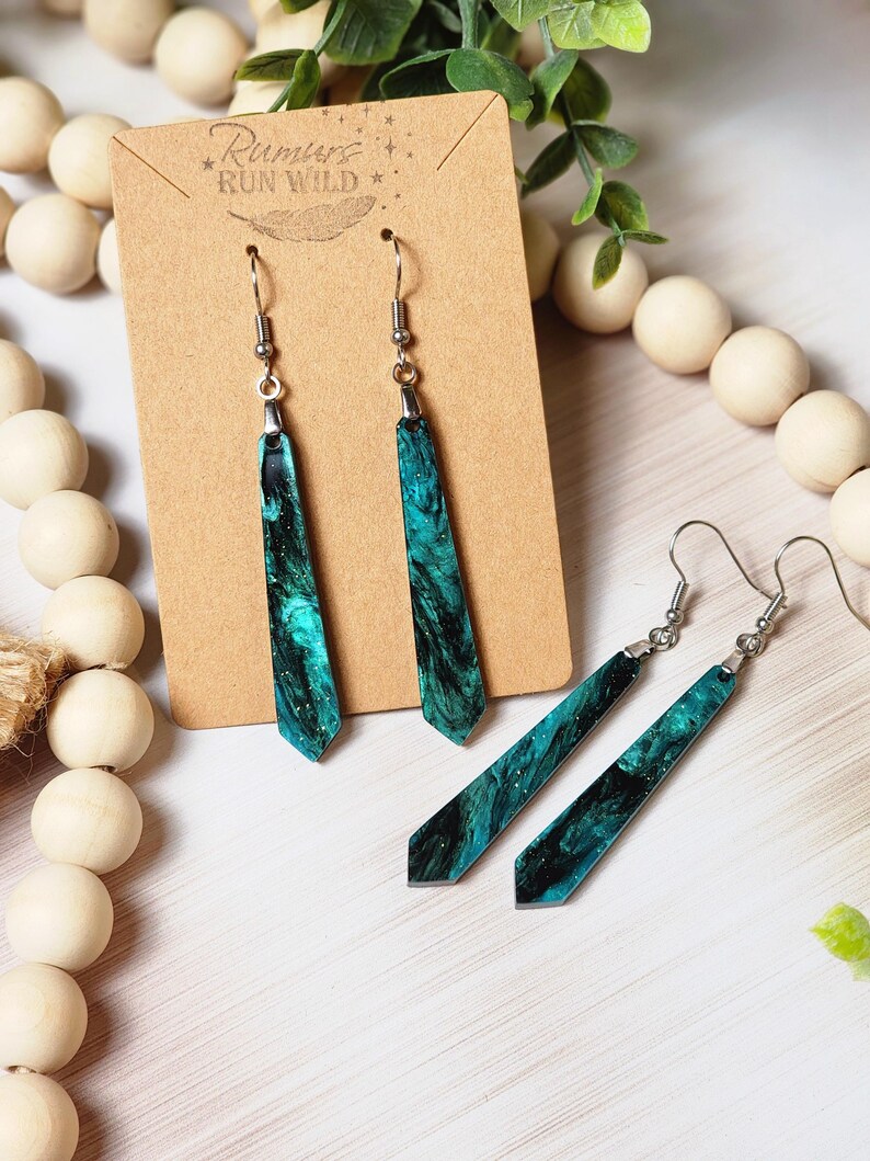 May include: A pair of long, teal and black dangle earrings with a silver hook. The earrings are made of resin and have a marbled effect. The earrings are on a brown card with the text "Rumours Run Wild" printed on it.