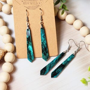 May include: A pair of long, teal and black dangle earrings with silver hooks. The earrings are made of resin and have a marbled effect. The earrings are displayed on a brown card with the text "Rumours Run Wild" printed on it.