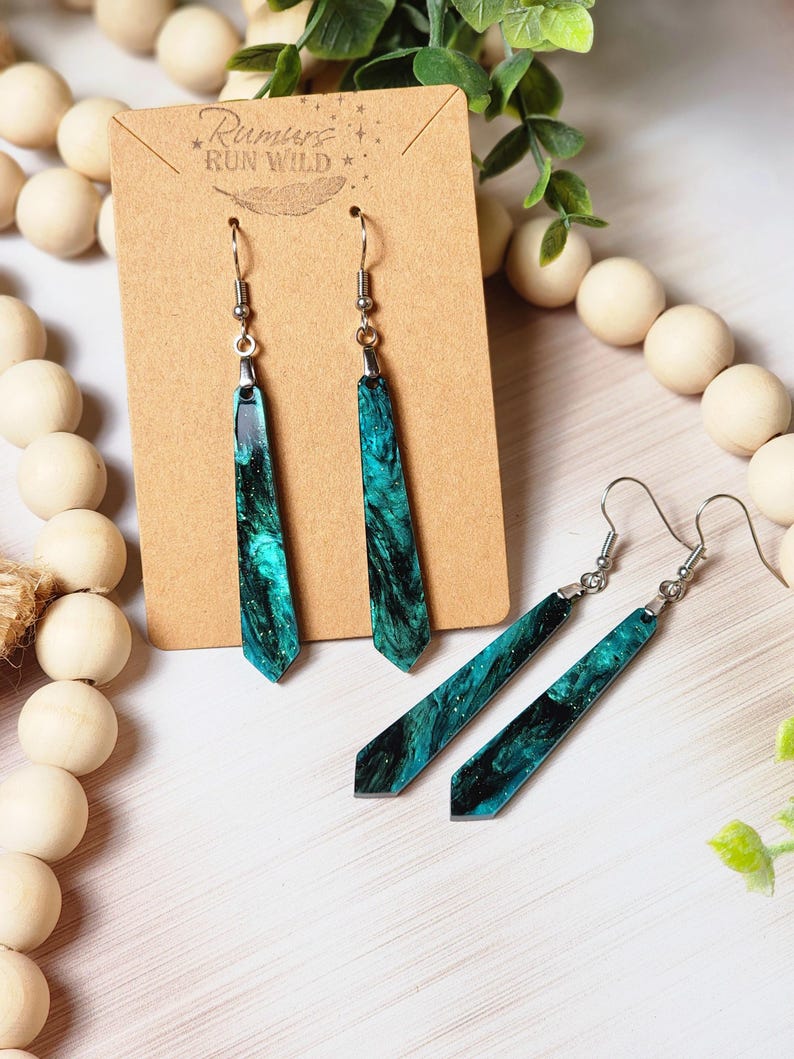 May include: A pair of long, teal and black dangle earrings with silver hooks. The earrings are made of resin and have a marbled effect. The earrings are displayed on a brown card with the text "Rumours Run Wild" printed on it.