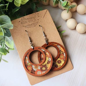 May include: Wooden hoop earrings with a floral pattern in shades of orange, yellow, and brown. The earrings are displayed on a brown card with the text "Rumurs Run Wild". The earrings have silver-colored ear wires.