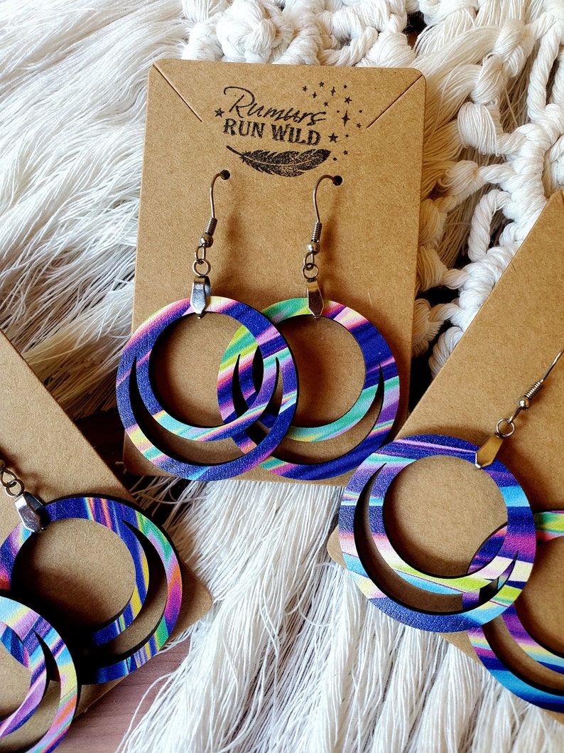 May include: A pair of wooden hoop earrings with a multi-colored swirl pattern. The earrings are on a brown card with the text "Rumours Run Wild" and a feather design.
