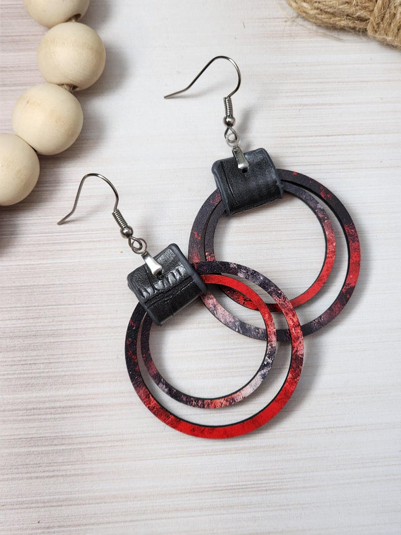 Puede incluir: Un par de pendientes colgantes con una parte superior cuadrada negra y tres aros circulares en capas. Los aros tienen un dise&ntilde;o veteado rojo, negro y blanco. Los pendientes tienen cierres de gancho de pez plateados.
