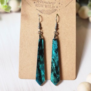 May include: A pair of teal blue and black resin dangle earrings with silver hooks. The earrings are shaped like a long, thin teardrop and have a sparkly finish. The earrings are on a brown card with the text "Rumors Run Wild" printed on it.