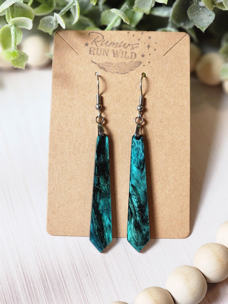 May include: A pair of teal blue and black resin dangle earrings with silver hooks. The earrings are shaped like a long, thin teardrop and have a sparkly finish. The earrings are on a brown card with the text "Rumors Run Wild" printed on it.