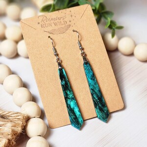 May include: Teal and black resin dangle earrings with silver hooks. The earrings are on a brown card with the text "Rumors Run Wild" printed on it.