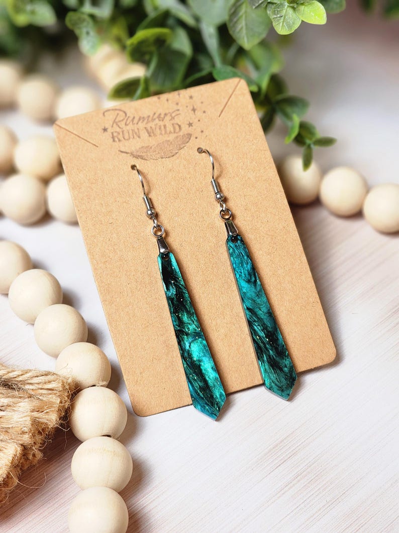 May include: Teal and black resin dangle earrings with silver hooks. The earrings are on a brown card with the text "Rumors Run Wild" printed on it.