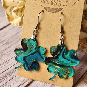 May include: A pair of green and gold shamrock earrings with a marbled effect. The earrings are on a brown card with the text "Run Wild" printed on it.