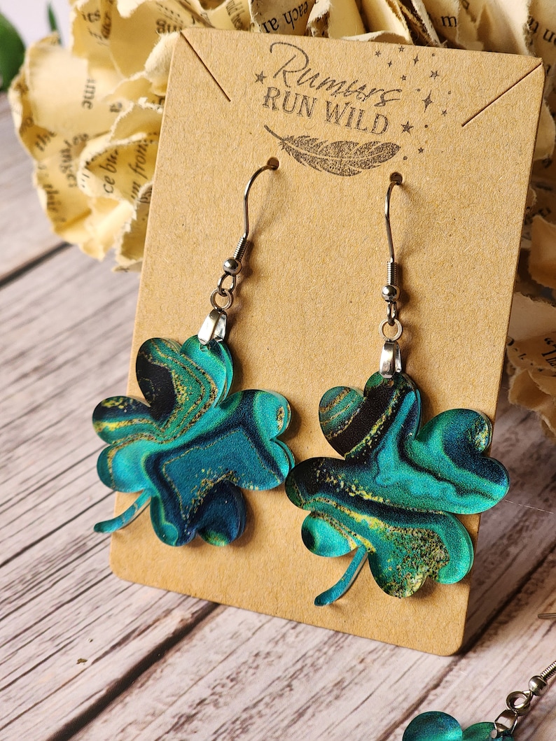 May include: A pair of green and gold shamrock earrings with a marbled effect. The earrings are on a brown card with the text "Run Wild" printed on it.