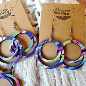 May include: A pair of wooden hoop earrings with a layered design. The earrings are painted with a colorful abstract pattern in shades of blue, green, pink, and white. The earrings are hanging on a brown card with the text "Rumors Run Wild" printed on it.