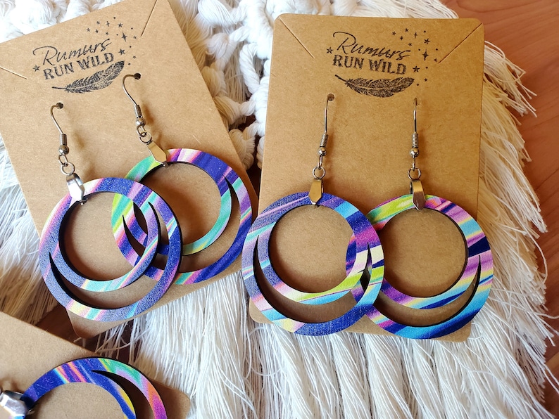 May include: A pair of wooden hoop earrings with a layered design. The earrings are painted with a colorful abstract pattern in shades of blue, green, pink, and white. The earrings are hanging on a brown card with the text "Rumors Run Wild" printed on it.