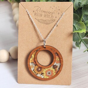May include: Wooden pendant necklace with a silver chain. The pendant is a double circle design with a floral pattern in yellow, orange, and white. The necklace is displayed on a brown card with the text "Rumors Run Wild".