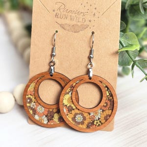 May include: Wooden hoop earrings with a floral pattern in shades of yellow, orange, and brown. The earrings have a circular design with a smaller inner circle. The earrings are displayed on a brown card with the text "Rumors Run Wild".