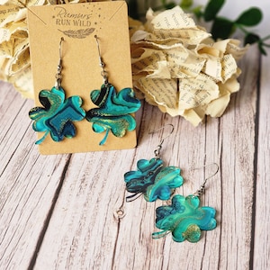 May include: A pair of dangle earrings featuring a four-leaf clover design. The clovers are made of resin and have a marbled blue and green pattern with gold glitter accents. The earrings are hanging from silver hooks.