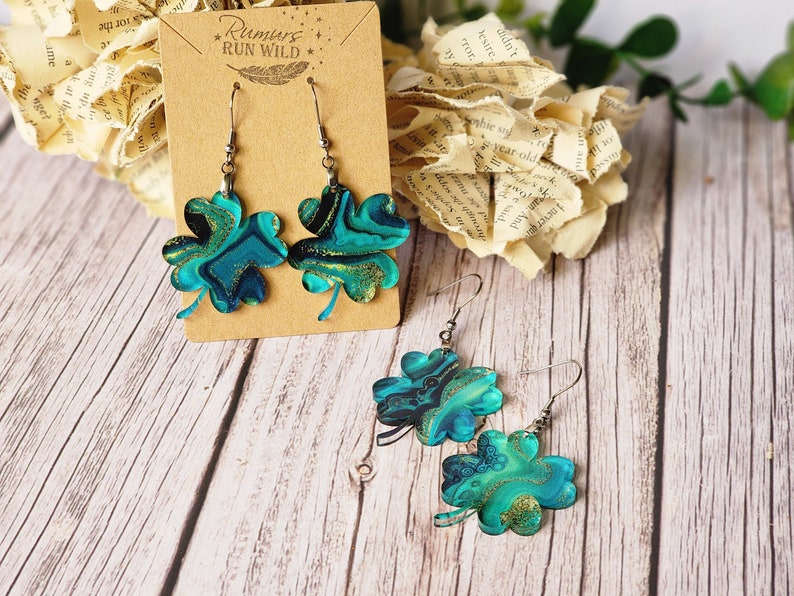 May include: A pair of dangle earrings featuring a four-leaf clover design. The clovers are made of resin and have a marbled blue and green pattern with gold glitter accents. The earrings are hanging from silver hooks.