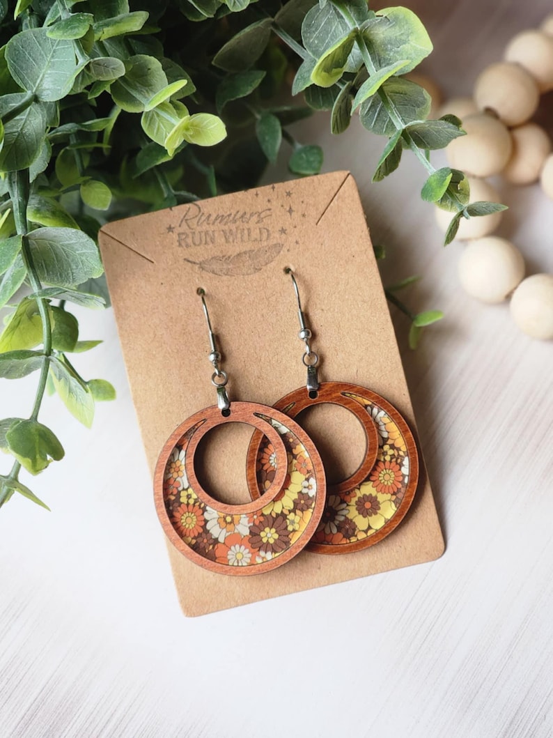 May include: Wooden hoop earrings with a retro floral pattern in shades of orange, yellow, and white. The earrings are on a brown card with the text "Rumurs Run Wild". The earrings have silver-colored hooks.